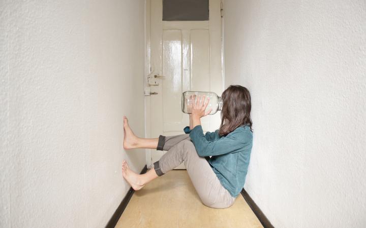 Eine Person sitzt in einem schmalen Gang auf dem Boden mit den Füßen an der Wand und hält sich ein leeres Glasgefäß an das Gesicht. 