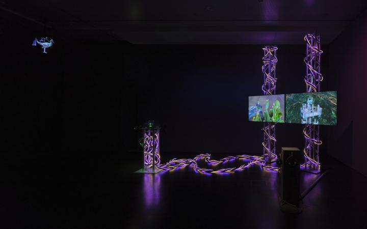 Auf dem Bild ist ein dunkler Raum zu sehen. Rechts im Bild befindet sich ein Kunstwerk/ Installation, bestehend aus zwei Bildschirmen, die von Leuchtelementen umgeben sind.