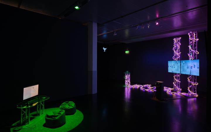 Auf dem Bild ist ein dunkler Raum zu sehen, in dessen Mitte sich zwei Kunstwerke befinden. Links ist ein grün ausgeleuchtet Platz mit einem Computer und zwei Sitzsäcken und rechts befindet sich eine Installation mit zwei Bildschirmen.