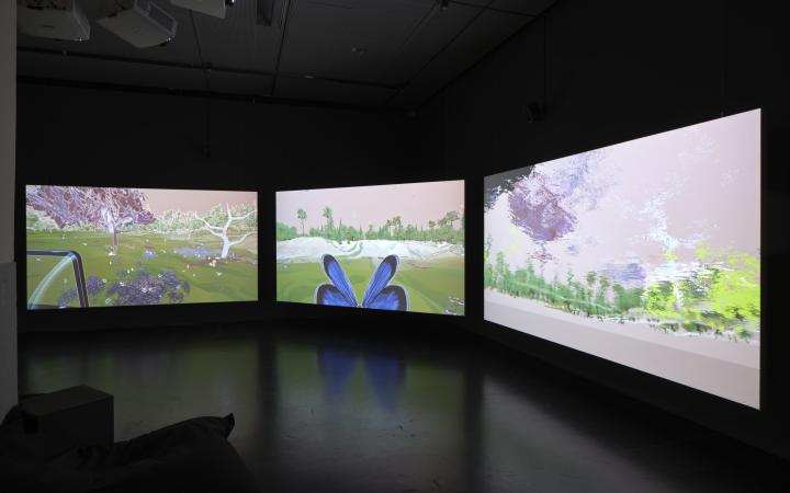 Auf dem Bild ist ein dunkler Raum zu sehen, in dessen Mitte drei große Bildschirme aufgestellt sind. Die Bildschirme zeigen eine Ki-generierte schöne Landschaft mit viel grün und einem Schmetterling.