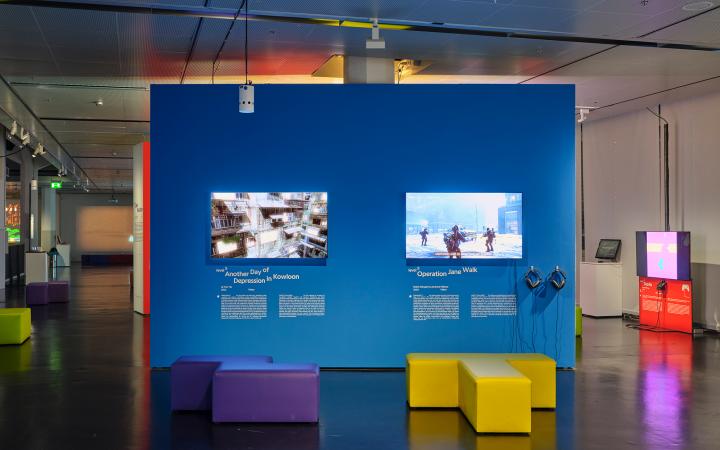 Blaue, freistehende Wand in der Ausstellung »Playful Architectures«. Daran angebracht sind zwei Bildschirme, die Computerspiel zeigen. Im restlichen Ausstellungsraum verteilen sich weitere Bildschirme.