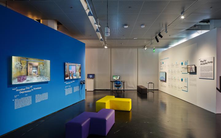 Blaue Wand in der Ausstellung »Playful Architectures« auf der linken Seite. Daran angebracht sind zwei Bildschirme, die Computerspiel zeigen. Im restlichen Ausstellungsraum verteilen sich weitere Bildschirme.