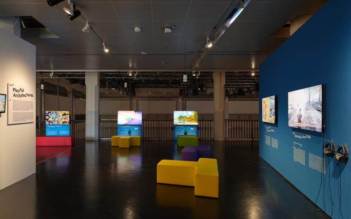 Blaue Wand in der Ausstellung »Playful Architectures« auf der rechten Seite. Daran angebracht sind zwei Bildschirme, die Computerspiel zeigen. Im hinteren Ausstellungsbereich verorten sich weitere Bildschirme. Links steht eine weitere Ausstellungswand.