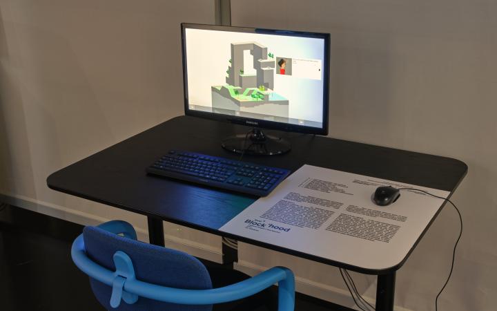 Ein blauer Stuhl steht vor einem Schreibtisch. Auf diesem Tisch steht ein Computerbildschirm mit angeschlossener Tastatur und Maus. Das Computerspiel »Block’hood« wird auf dem Bildschirm ausgespielt.