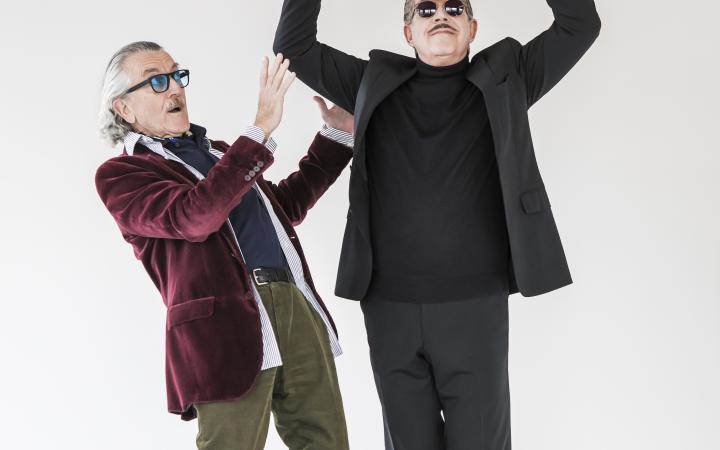 The musician duo Yello stands side by side in front of a wall. The one on the right wears a suit and holds a sculpture of two round balls above his head. The left one stands next to him in an intercepting position.
