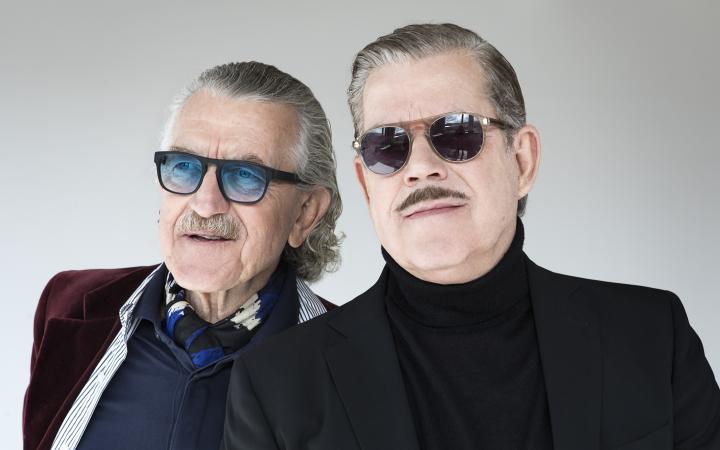 Zwei Männer stehen nebeneinander, die beiden Köpfe sind groß zu sehen. Der rechte trägt eine Sonnenbrille, einen Oberlippenbart, kurze, nach hinten gekämmte Haare und einen Rollkragen mit Jacket. Der linke trägt eine gefärbte Sonnenbrille.
