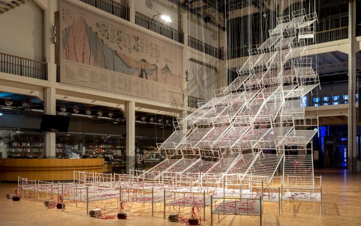 Installation view of Chiaru Shiota's »Connected to Life«. Hospital bed frames can be seen hanging from the ceiling in the shape of a pyramid. These are crisscrossed by tubes through which red paint flows.