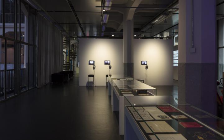 In the background there is a white wall with small screens and headphones. In front of them black bar stools. In the middle and foreground are three vitrine tables with books.