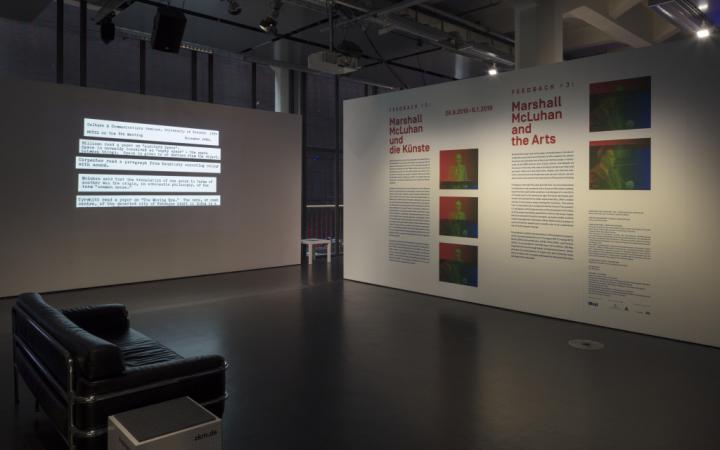 In the foreground on the left side is a black sofa. At the back left there is a partition wall with text projection. In the back on the right there is a wall printed with text and pictures.