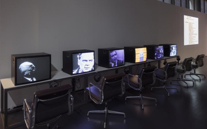 On a wall, a long table with six televisions on which different videos run. Headphones and black chairs in front of them.