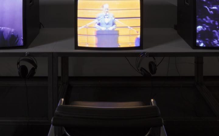 Close-up of a video installation. Television on table with headphones and a black chair in front of it.