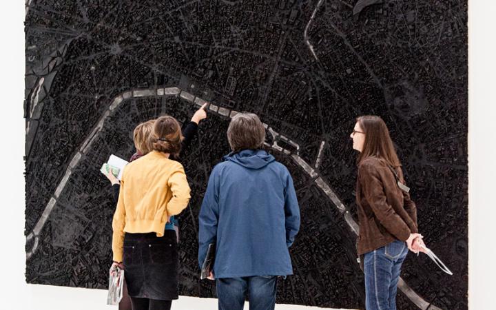 tTo see are four people who are in front of a three-dimensional picture hanging on the wall. The image resembles a city map, which is entirely colored in black and crossed by a few white lines