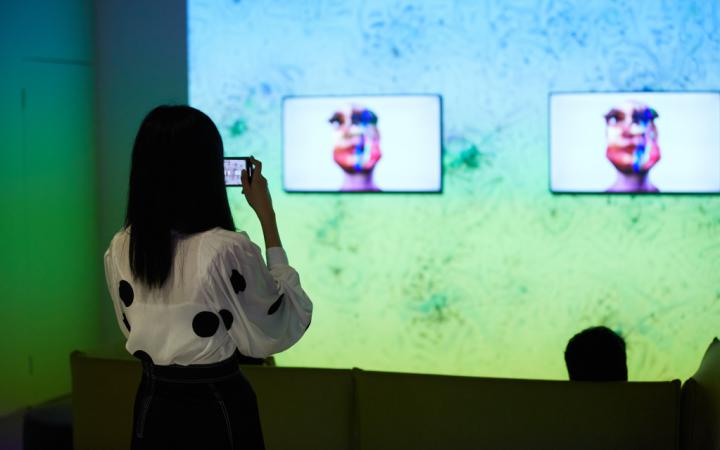 The photo shows the work »im here to learn so :)))))))«. In the background is a large screen on which a Google DeepDream is projected. Three screens in front of it show the head of the chatbot Tay.