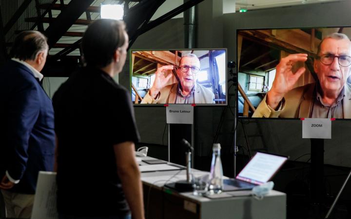 Bruno Latour ist in zwei Bildschirmen zu sehen. Zwei Männer, einer davon Peter Weibel, stehen vor den beiden Monitoren.