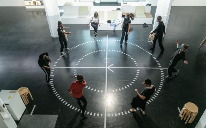 Eine Gruppe Menschen steht im Kreis auf einem runden Diagramm auf einem Hallenboden. Die Menschen scheinen in Bewegung zu sein.