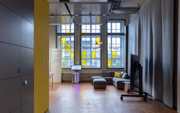 Blick in die Ausstellung auf eine gelbe Wand, eine Couch und das Werk "Eye-Tracking-Kiosk" bestehend aus einem Bildschirm und einem davonstehlenden Hocker