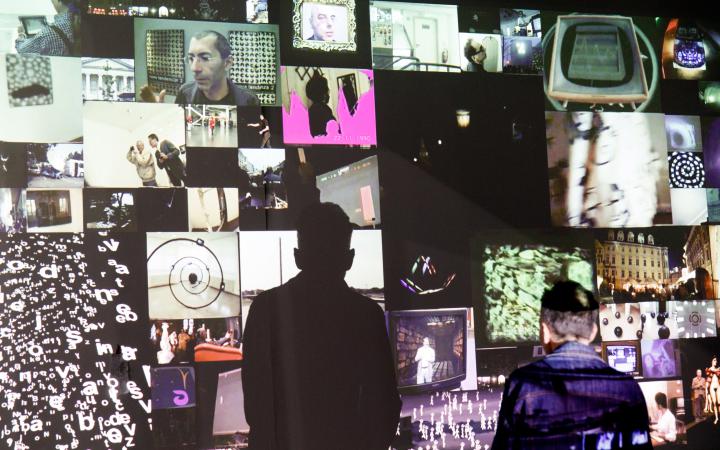 You can see a video installation. Many smaller images are projected onto a wall, creating a mosaic-like image. In front of it stands a man. He casts a conspicuous shadow on the wall.
