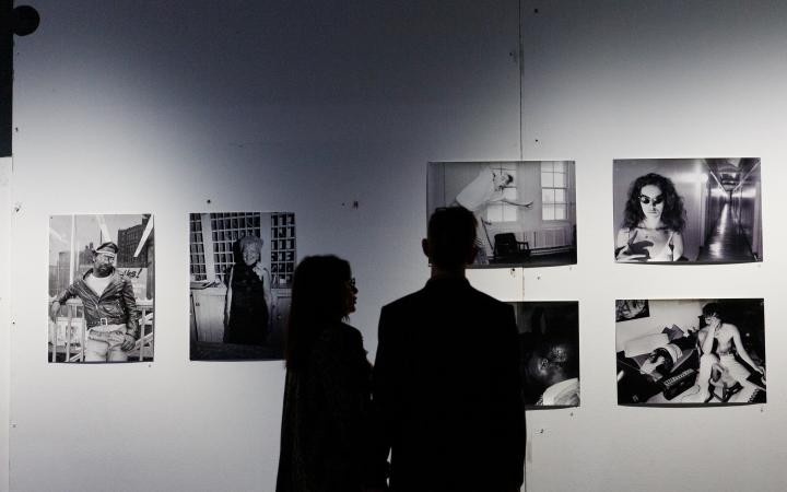 Zwei Personen, die nur als Schatten zu erkennen sind, stehen vor einer Wand mit mehreren Schwarz-Weiß-Fotografien.