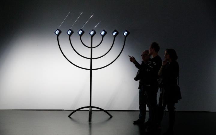 Zu sehen ist eine große, schwarze Menorah, die anstelle von Kerzen sieben kleine Bildschirme, mit nach rechts gerichteten Antennen, hat. Auf der rechten Seite stehen drei Personen, die sie betrachten. Eine der Personen zeigt auf die Installation.