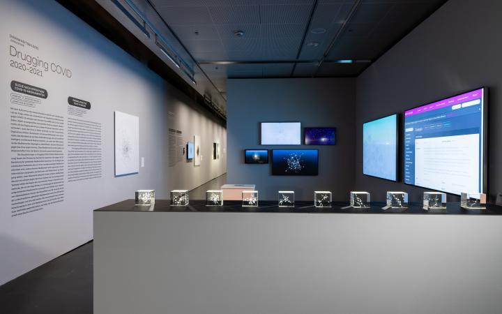An exhibition space. In the foreground are 10 small glass boxes showing fine networks. In the background hang 4 screens that also show networks. On the right hang two large screens, on the left is an explanatory text on the wall.