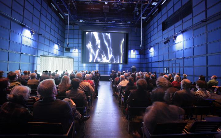 Das ZKM_Medientheater voller Menschen, die auf eine Leinwand schauen, auf der amorphe Strukturen in weiß zu sehen sind
