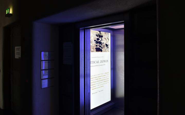 Zu sehen ist ein geöffneter Fahrstuhl. An der Innenwand des Fahrstuhls befindet sich ein großer Display, welcher einen Text zu der Ausstellung Critical Zone zeigt. 