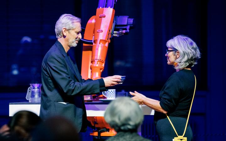 In the background is an orange robot that has just made tea, which the artistic and scientific director of the ZKM | Karlsruhe Alistair Hudson is handing to a woman.