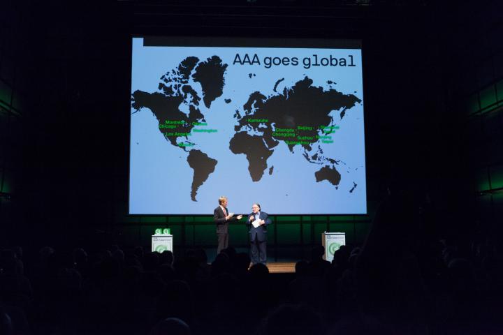 Zwei Männer auf der Bühne. Im Hintergrund eine Weltkarte mit »AAA goes global«