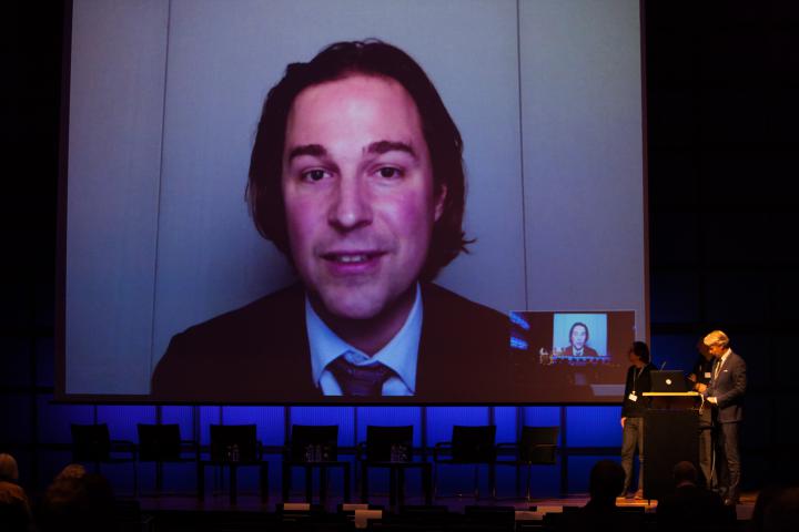 Ein sprechender Mann auf einem Screen sowie drei Männer auf einer Bühne.