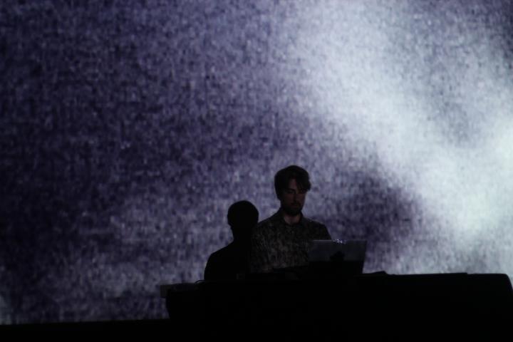 Mann auf der Bühne vor einem Laptop. Hintergrund ist in weißem Licht getaucht