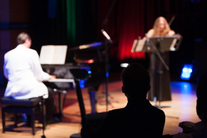 A man in a white suite playing piano. A woman in a black dress playing violin.