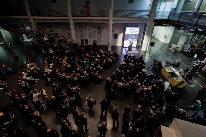 View from above: people standing in front of a stage on which a man makes a speech.