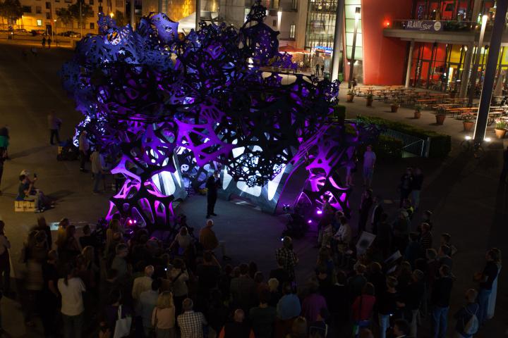 A black construction dipped in purple light. In the midst of the construction people are standing.