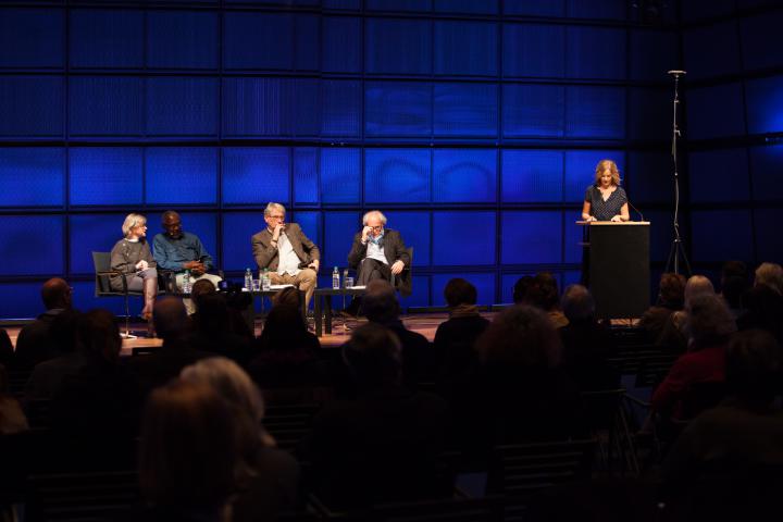 Vier Personen sitzen auf einer Bühne. Eine weitere Person steht rechts von ihnen an einem Podest und spricht zu dem Publikum