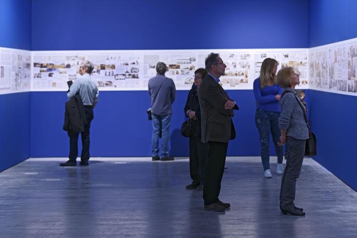 Menschen sehen sich Bilder der Ausstellung an, die auf blauem Hintergrund ausgestellt sind