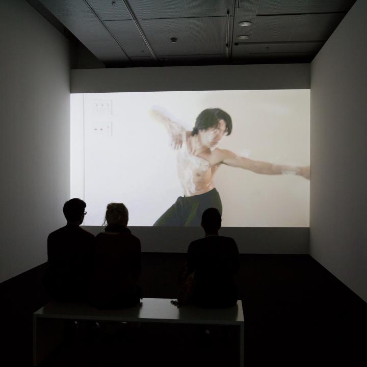 Three persons sit in front of a screen on which a man, wrapped in cling film, can be seen