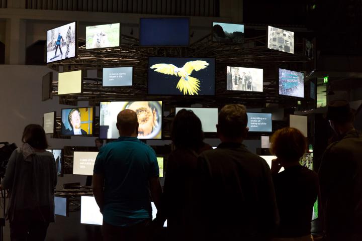 Das Bild zeigt BesucherInnen vor einer kreisförmig angeordneten Vielzahl von unterschiedlich bespielten Bildschirmen. 