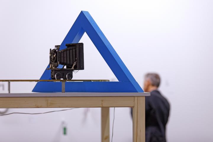 Das Bild zeigt eine Kamera, die durch ein hohles, blaues Dreieck gerichtet ist. 