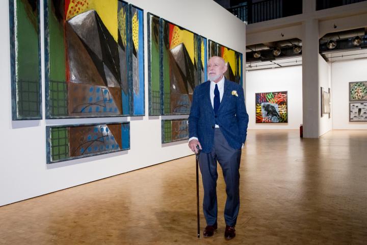 Markus Lüpertz steht im Anzug vor einem bunten Bild in der Ausstellung