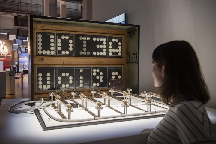 A woman stands in front of an installation consisting of several interconnected parallel bars and a box displaying letters.