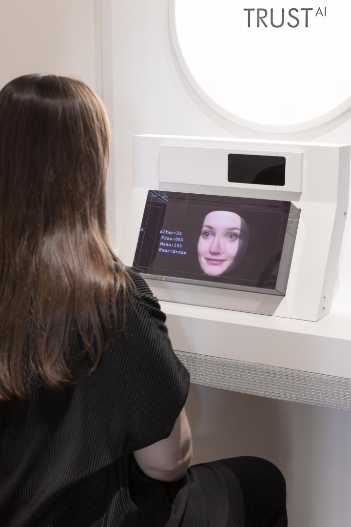 A visitor can be seen. She is seated with her back turned and is interacting with the media installation. She is talking to a female artificial intelligence that is facing her.
