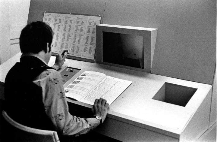A man sits at a large computer with a notebook and a table