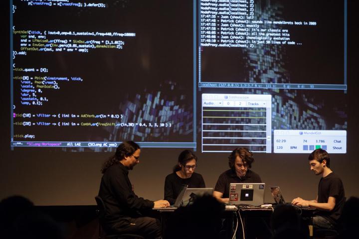 Four programmers sit in front of their computers while the produced data can be seen in the background on the screen