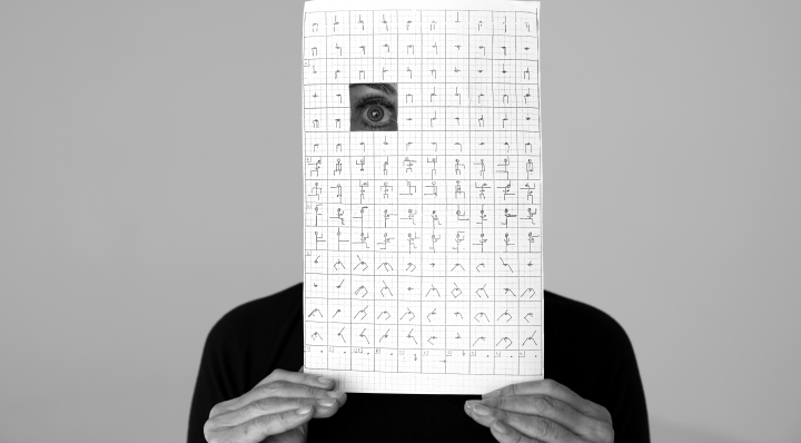 Analivia Cordairo can be seen. Her face is covered by a sheet of paper on which there are drawings of a choreography. Only one of her eyes is visible, as a square has been cut out of the paper.