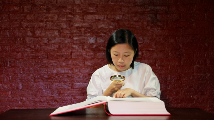 A woman reads a large open book in front of a red wall with a magnifying glass