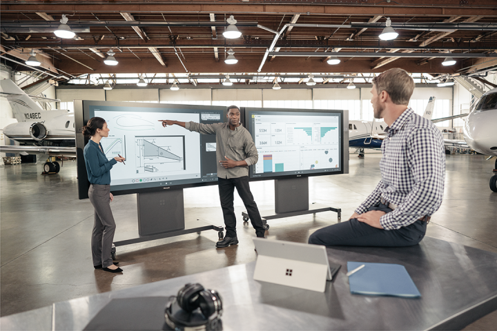 Drei Menschen arbeiten an digitalen Bildschirmen in einer Halle mit Flugzeugen