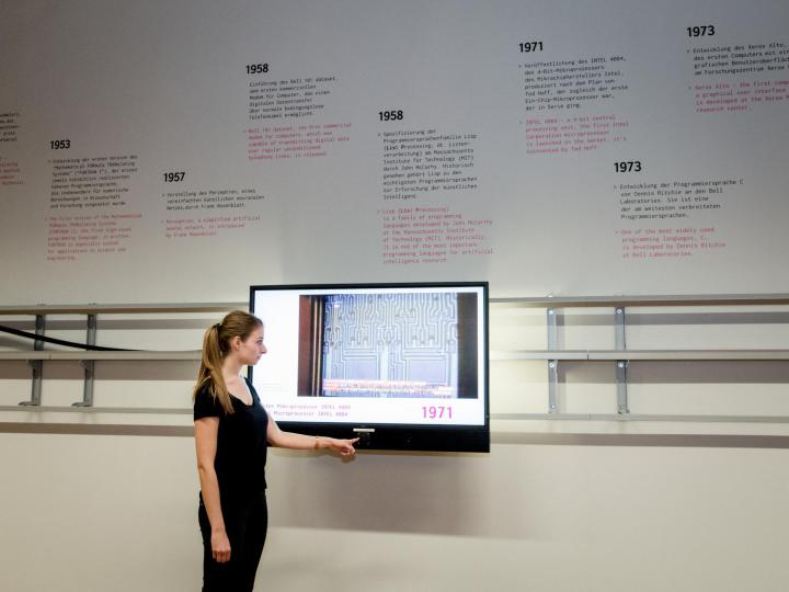Das Bild zeigt eine Frau im Halbprofil, die die virtuelle Zeitleiste der »Genalogie des digitalen Codes« bedient. An der Wand, wo der Bildschirm auf einer beweglichen Schiene befestigt ist, ist eine Infografik zu sehen