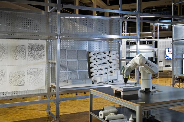Im Vordergrund steht ein Roboterarm auf einem Holztisch, während man im Hintergrund verschiedene Plastiken sowie Zeichnungen an einer Gitterstruktur sieht