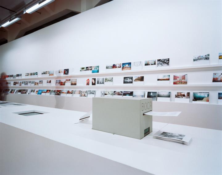 The picture shows a shelf with three levels where photographs are displayed. In the foreground you can see a photo printing machine