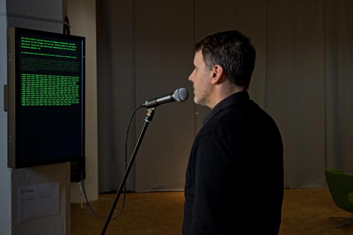 Ein ins Mikrofon sprechender Mann steht vor einem Bildschirm auf dem ein deutscher Text.  Morseschrift und Computersprache zu sehen sind